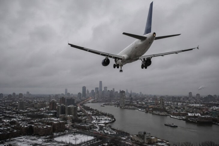 An Airbus A320 aircraft in low-altitude flight over an urban area and river, shot from below in overcast winter light