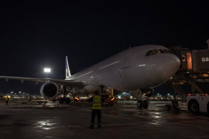 An Airbus A330 aircraft at a terminal gate at night, illuminated by ground floodlights during a late-night departure
