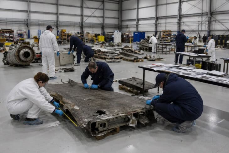 Aviation accident investigators examining recovered aircraft components in a hangar, working methodically with documentation at hand