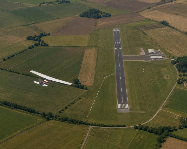 Aerial view of a light aircraft in the traffic pattern at a small airfield, showing the base leg and the turn onto final approach with the runway visible below