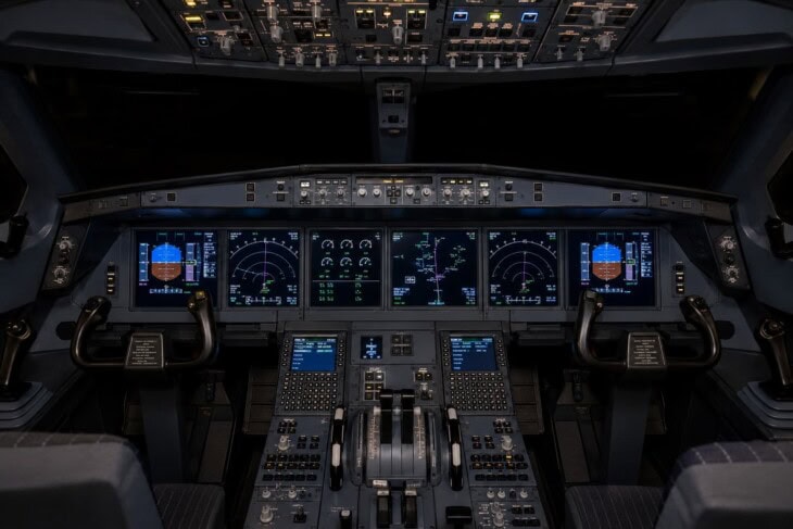 Modern commercial airliner glass cockpit flight deck showing multiple large LCD screens with primary flight displays, engine indication screens, and navigation displays
