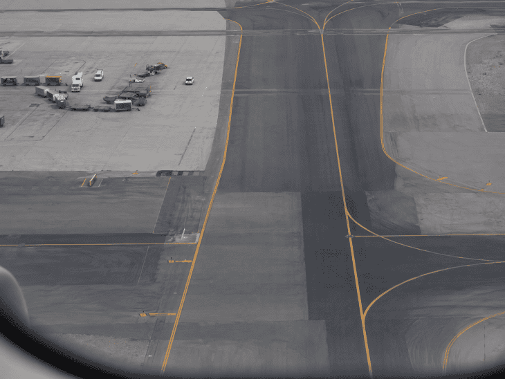 Airport tarmac lines and markings viewed from an aircraft window during taxi, yellow and white markings crossing dark grey pavement, service vehicles as small colorful shapes against the geometric pattern