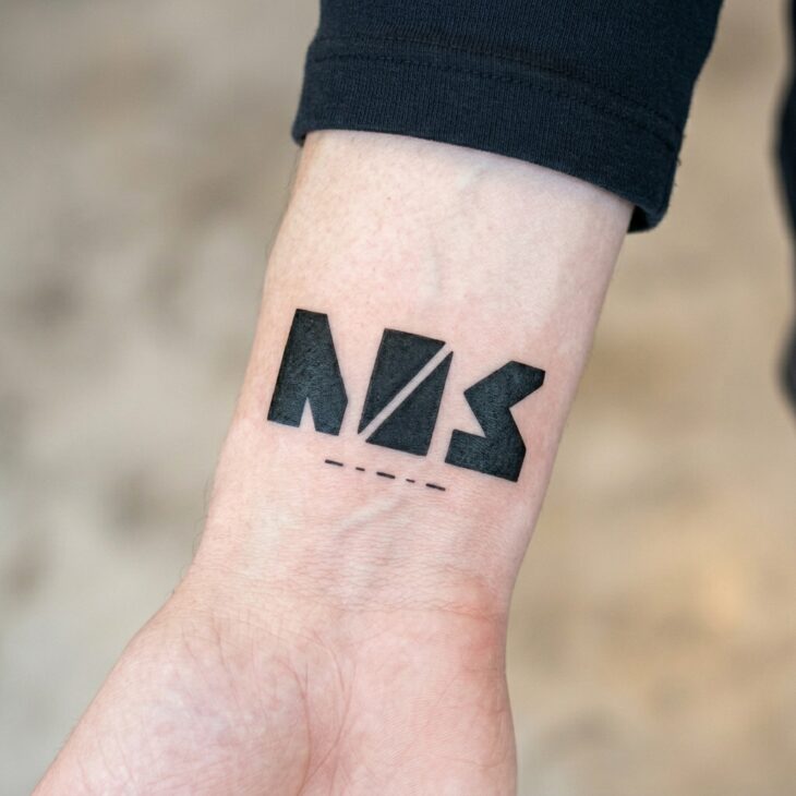 Minimalist airport-code style tattoo on an adult wrist with three block letters and a small runway line beneath