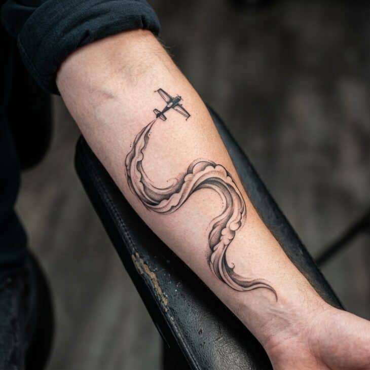 Dynamic airshow smoke trail tattoo along an adult outer forearm with a tiny generic aerobatic plane and sweeping shaded smoke ribbon