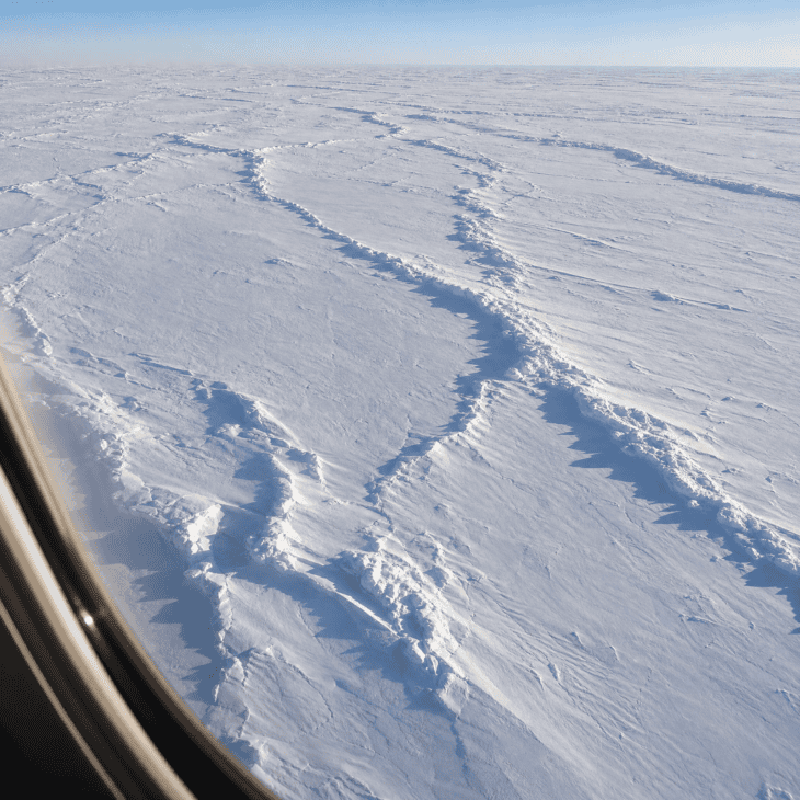 A vast Arctic ice field viewed from an aircraft window, broken by pressure ridges casting dark linear shadows across the white surface, horizon a flat white line meeting pale blue sky