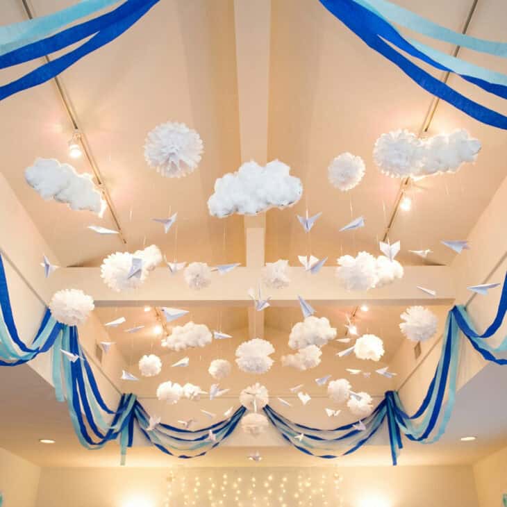 Party room ceiling decorated with white tissue pom-pom clouds and hanging origami paper planes on fishing line