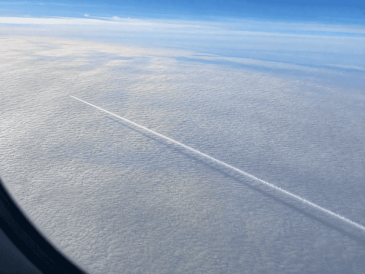 A white contrail stretches across a cloud layer below, its shadow visible as a thin dark line on the cloud surface when viewed from an aircraft window above