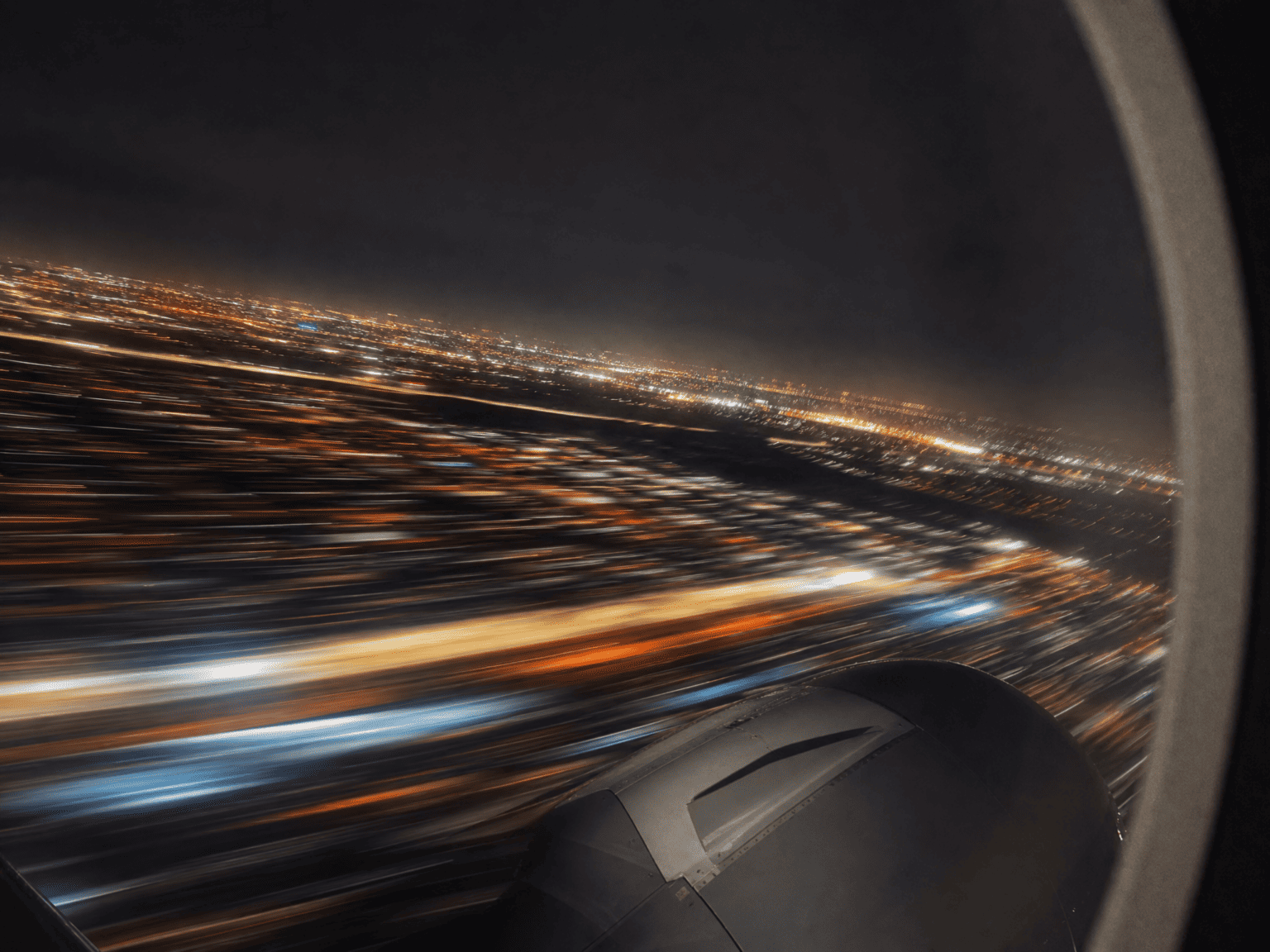 City lights streaked into amber and white motion trails through an aircraft window during a nighttime departure, the long-exposure blur giving the lights a painterly kinetic quality