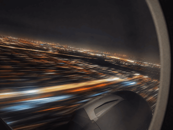 City lights streaked into amber and white motion trails through an aircraft window during a nighttime departure, the long-exposure blur giving the lights a painterly kinetic quality