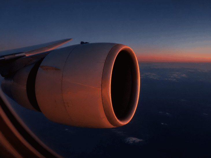 An aircraft engine photographed at dusk from the window seat just behind the wing, the nacelle catching the last amber light of day, the intake a dark oval, deep blue twilight sky behind it
