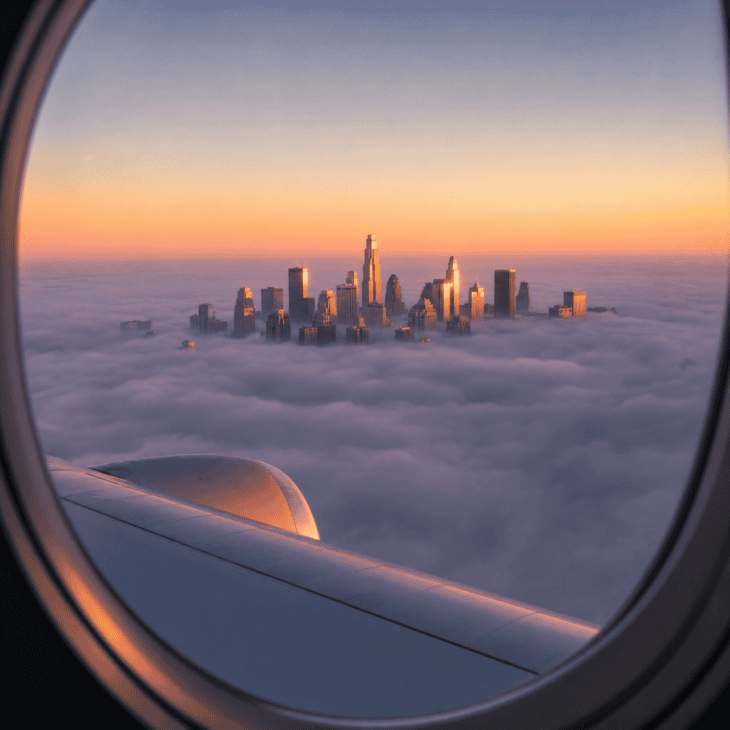 A city partially submerged in morning fog, the tallest buildings poking through the fog layer into warm peach sunrise light, the fog surface a cool lavender-grey below them
