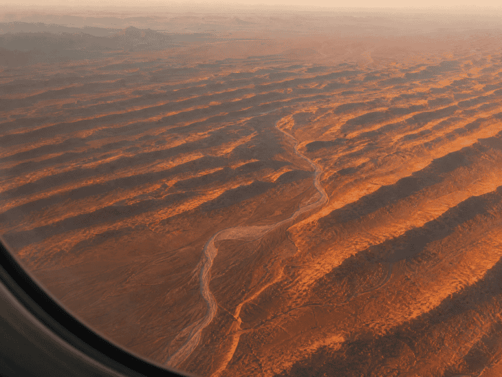 A desert landscape glowing amber and burnt orange in low afternoon sun, dune ridges casting long dark parallel shadows, a dry river channel winding through the terrain below an aircraft window