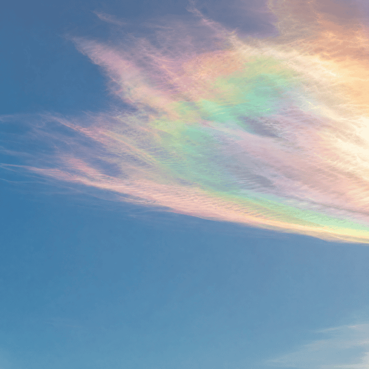 A thin high-altitude cloud near the sun displays shimmering iridescent bands of pink, green, violet, and gold caused by light diffracting through ice crystals, pale blue sky surrounding it