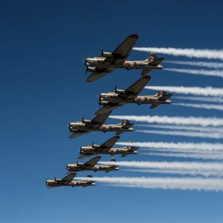 Formation of five WWII heavy bombers at high altitude with contrails streaming from all engines against a deep blue sky