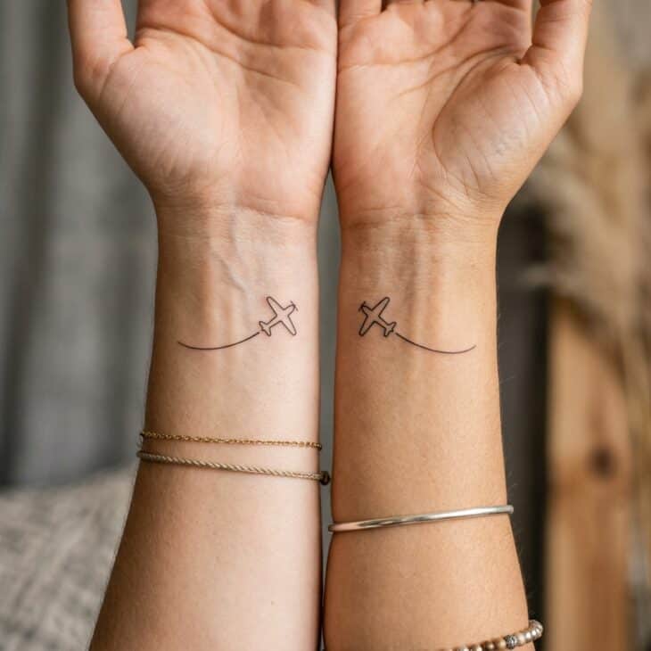 Two matching tiny fine-line airplane tattoos on two adult wrists with opposite curved flight trails