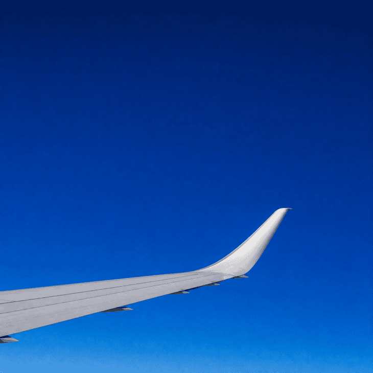 The curved tip of an aircraft wing against a deep, cloudless blue sky at cruise altitude, the wing surface a cool silver-grey, vast empty blue filling the upper frame