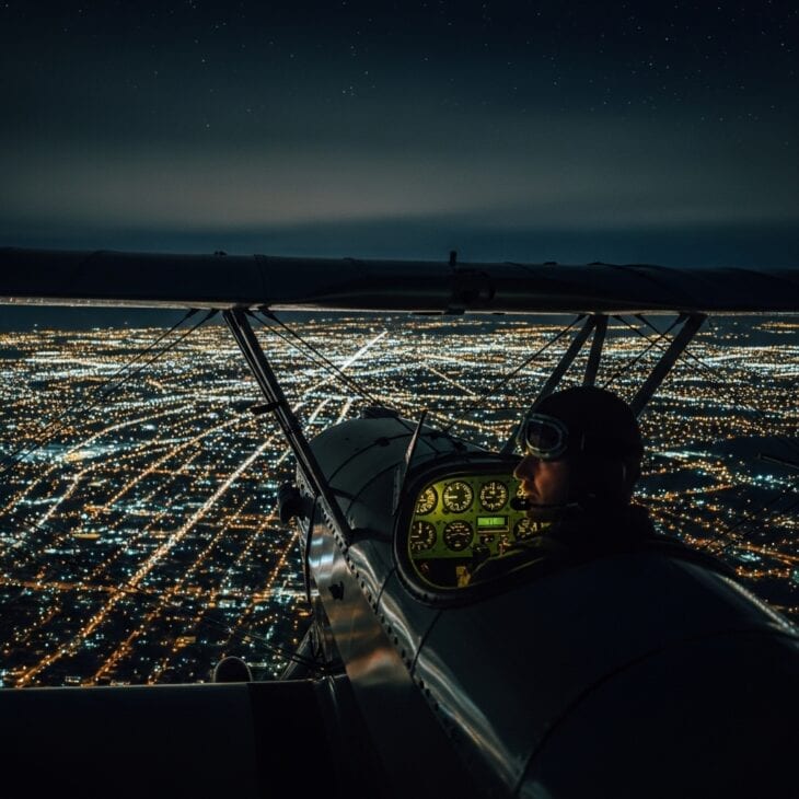 Open-cockpit pilot at night above a carpet of city lights, face illuminated by instrument glow against a dark sky with stars