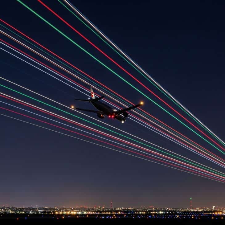 Long-exposure night shot of an aircraft on approach with navigation lights creating coloured light trails against a dark sky