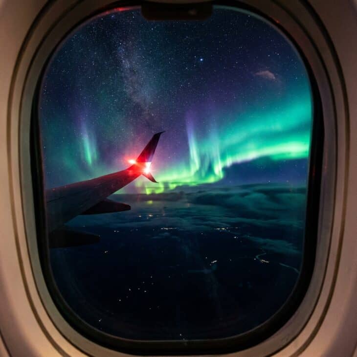 Northern lights visible as green and violet ribbons through an airliner window at night with the wing tip and nav light at frame edge