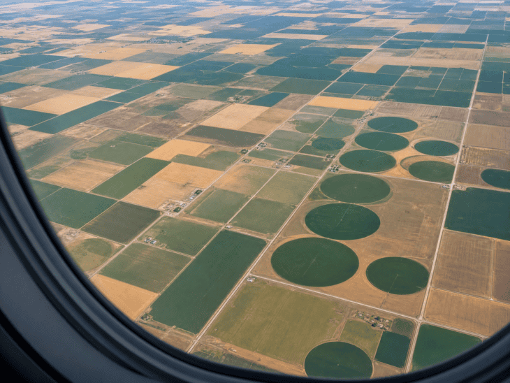 Agricultural fields viewed from an aircraft window form a patchwork of yellows, greens, and tans, with circular irrigation patterns creating green discs against bare earth