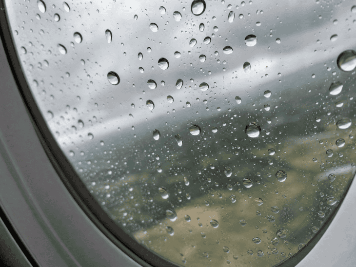 Water droplets of varying sizes cling to the exterior of an aircraft window during rain, each refracting a blurred green and gray landscape below