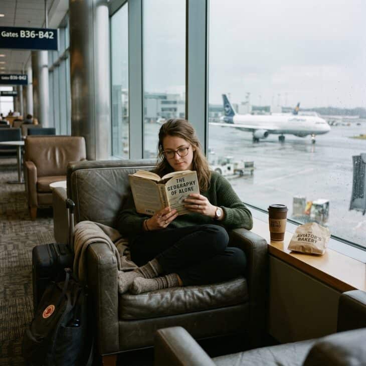 Traveler absorbed in a hardback book in a quiet airport lounge chair with a coffee and aircraft visible through the window