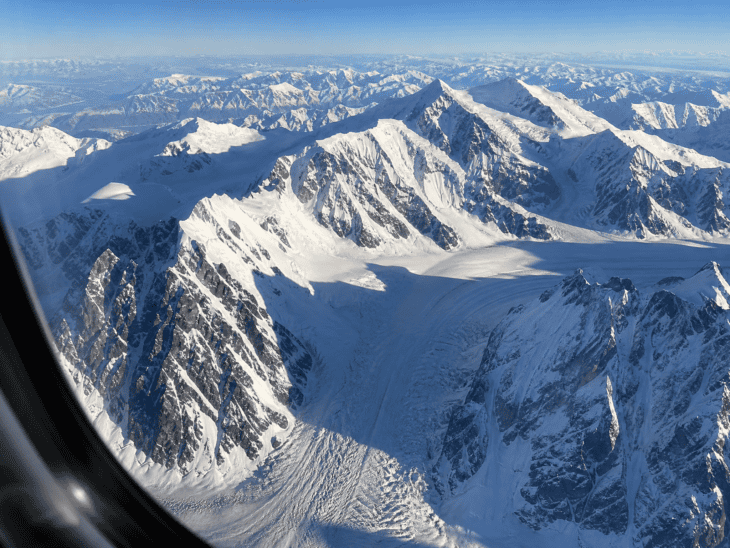 A snow-covered mountain range seen from altitude through an aircraft window, hard shadows from the peaks cutting across white snowfields, dark rock visible on steep faces