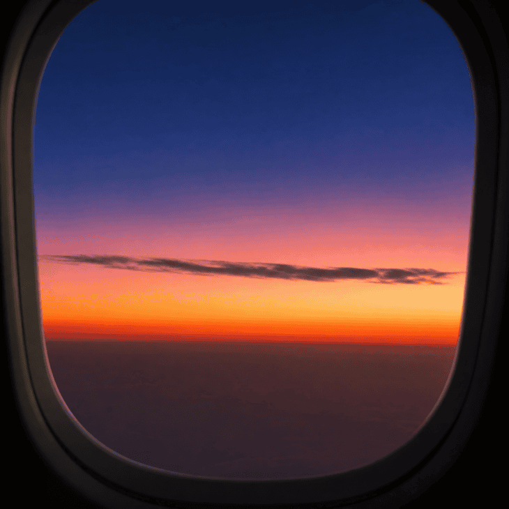 Stacked horizontal bands of sunset color viewed through an aircraft window: deep orange at the horizon graduating through rose, gold, lavender, and twilight blue, a single cloud silhouette crossing the gradient
