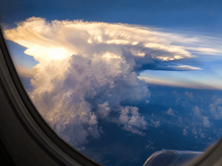A towering cumulonimbus cloud photographed from an aircraft window, the anvil top glowing brilliant white in sunlight with dark purple shadows in the cloud towers below