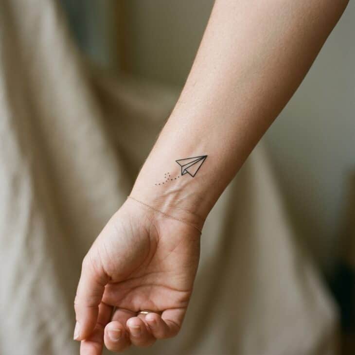 Small fine-line paper airplane tattoo on an adult inner wrist with a short dotted flight trail