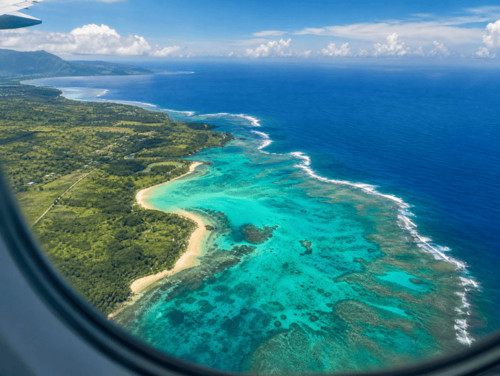 A tropical coastline seen from an aircraft window, water ranging from brilliant turquoise in the shallows to deep blue offshore, sandbars and reef formations visible below the clear surface