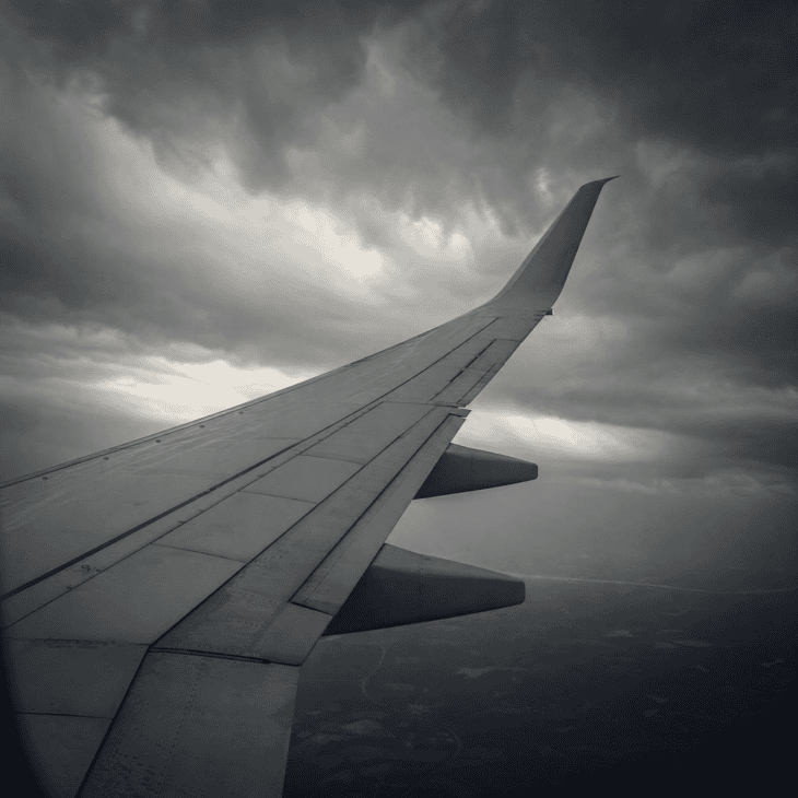 An aircraft wing flexed upward in a pronounced curve during turbulence, viewed from a window seat against a dark dramatic overcast sky, the wingtip clearly elevated above the wing root