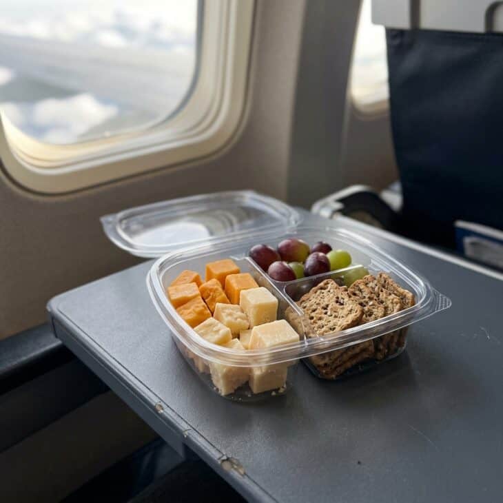 A clear snack container with cheese cubes, whole-grain crackers and grapes on an airplane tray table
