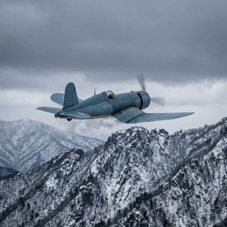 Gull-winged carrier fighter aircraft flying low over a snowy Korean mountain ridge