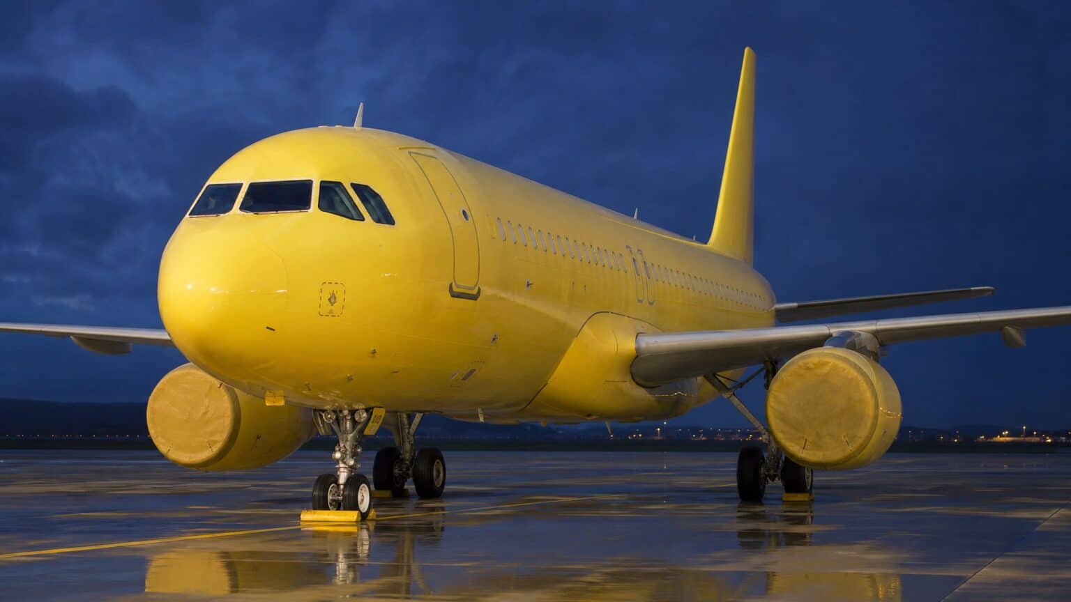 A yellow Airbus A320 parked at dusk with its engines covered and wheels chocked
