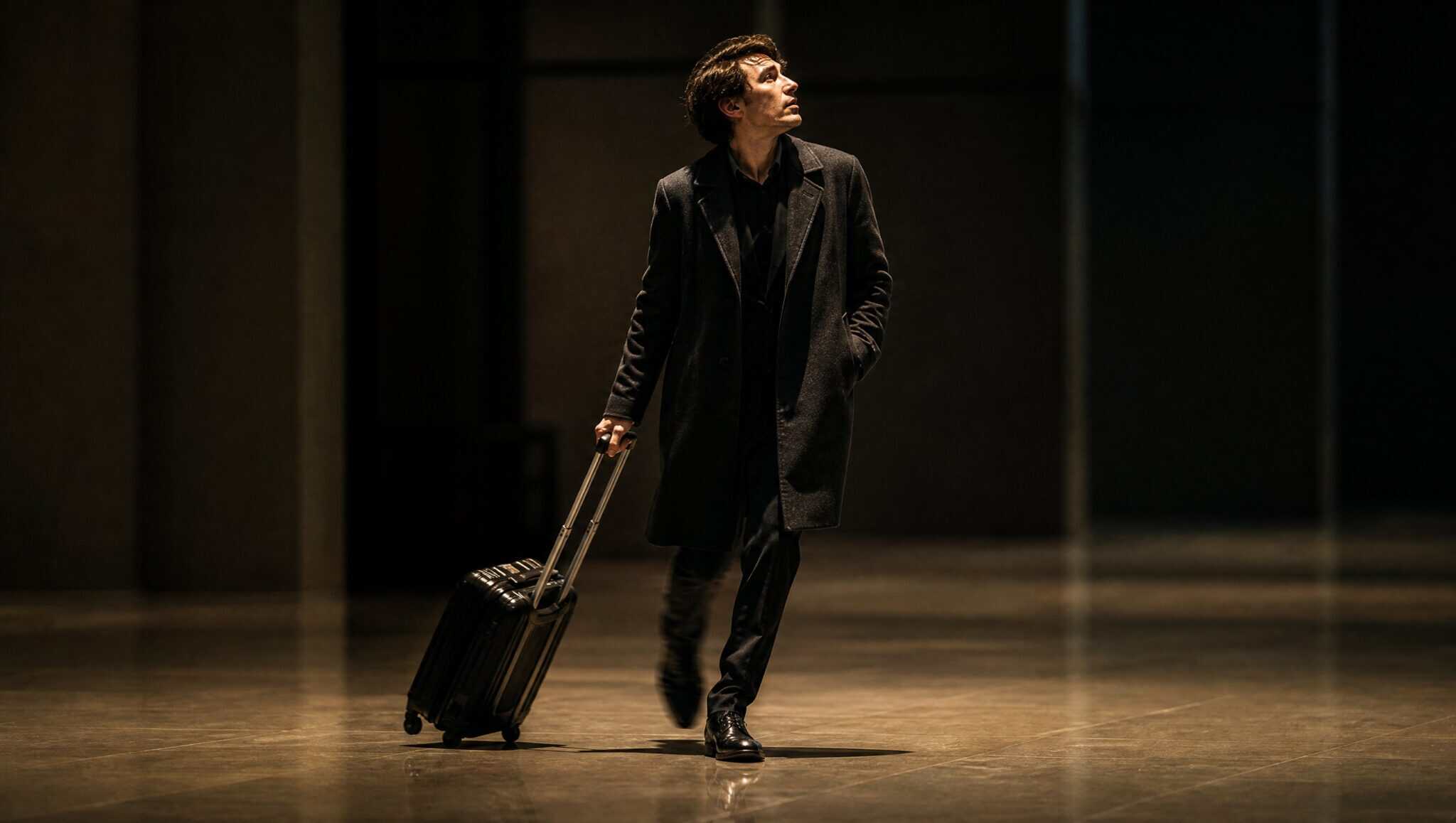 A lone traveller pulling a carry-on suitcase strides through a dark, empty airport terminal corridor
