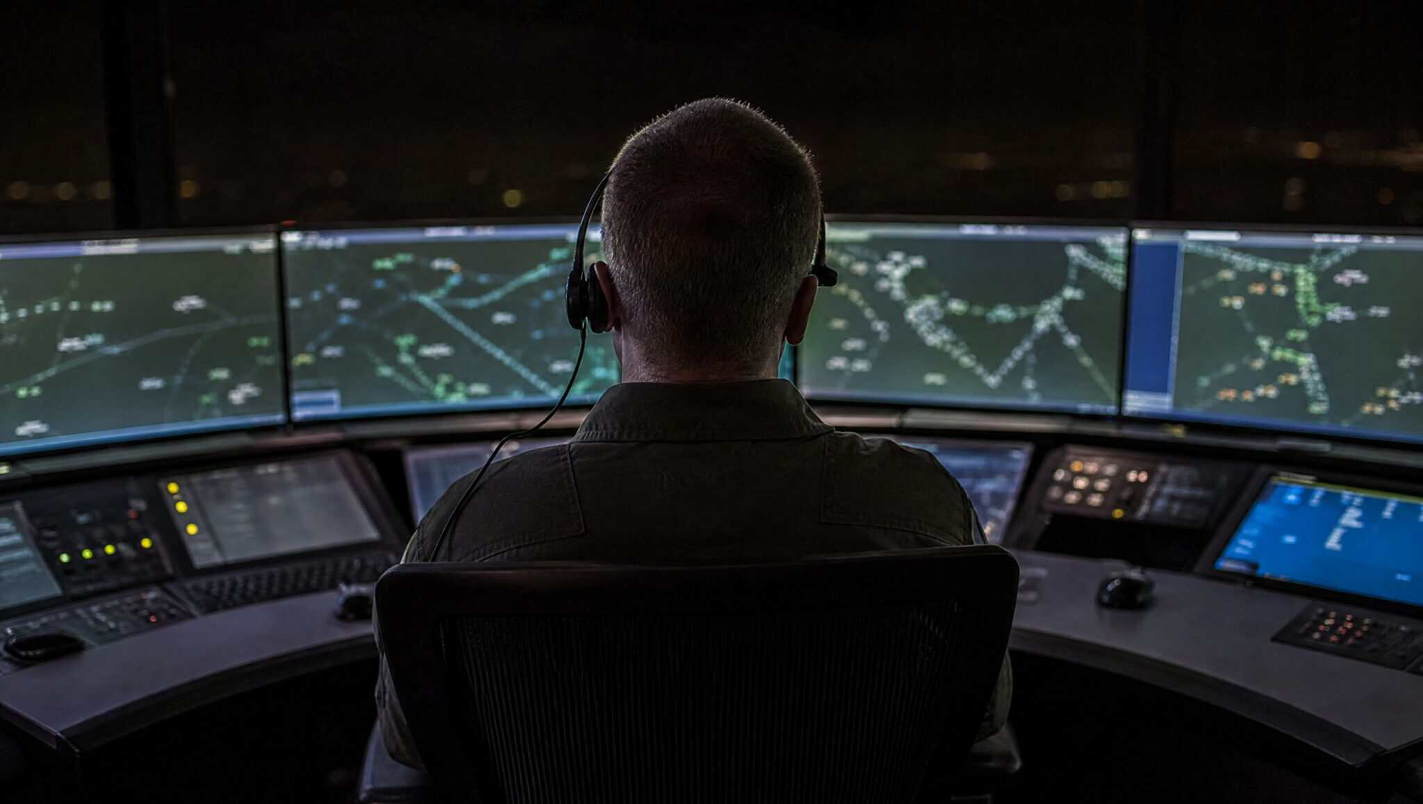 An air traffic controller seen from behind at a darkened en route control centre console, facing a curved bank of glowing radar displays