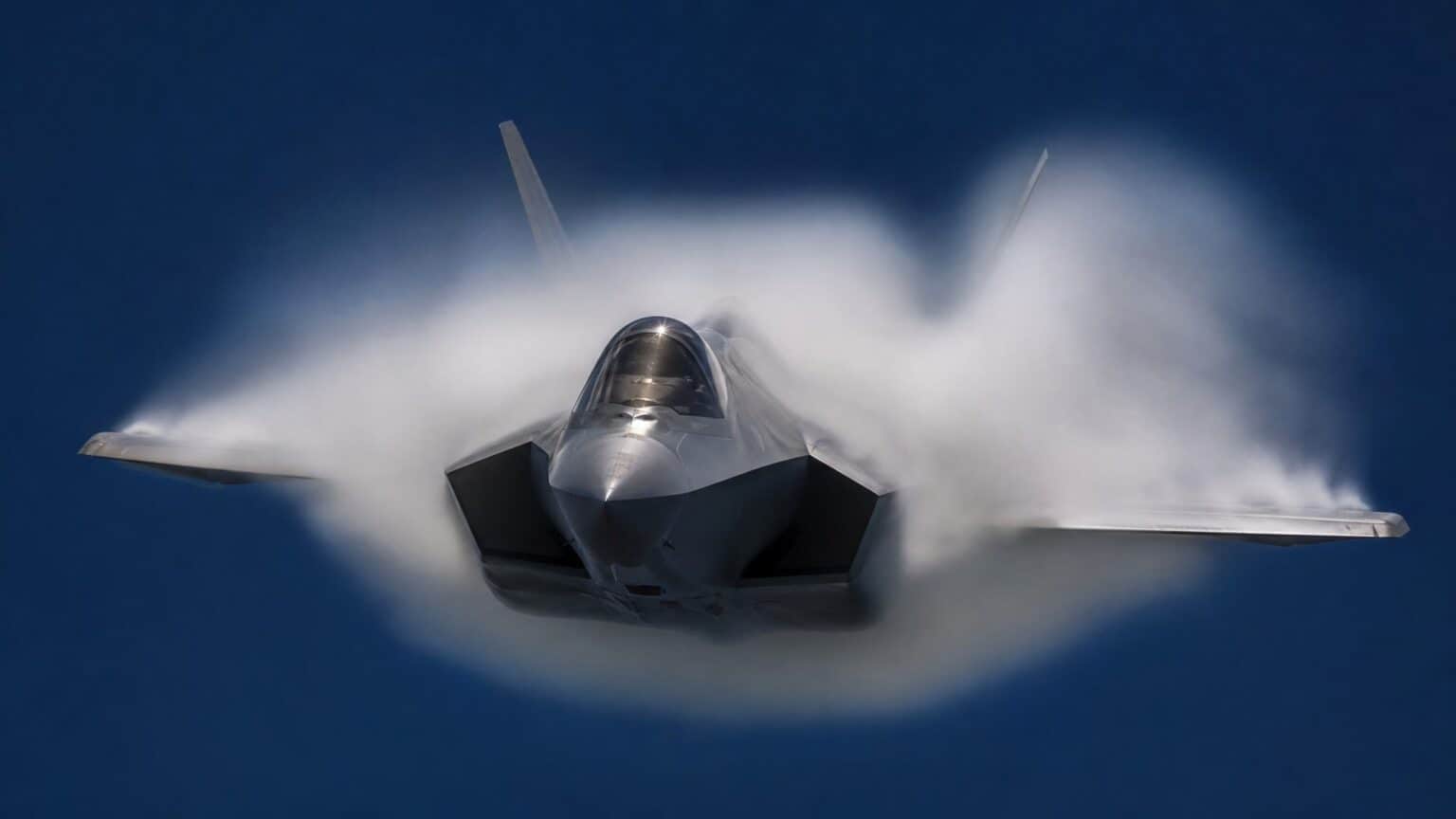A single-seat fighter jet wrapped in a white vapour cone as it accelerates through the transonic range against a dark blue sky