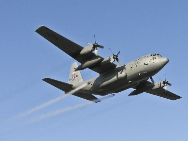 Gray Air Force Reserve C-130 Hercules seen from below against a blue sky, spray trailing from booms at its tail
