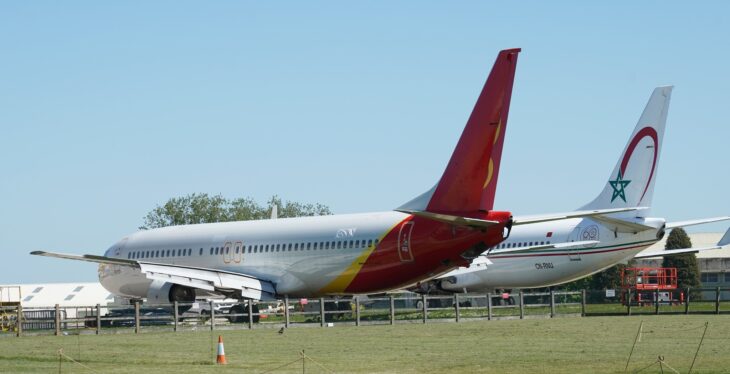 Two retired Boeing 737s, one with a red and yellow tail and one in Royal Air Maroc colors, parked behind a fence at Cotswold Airport