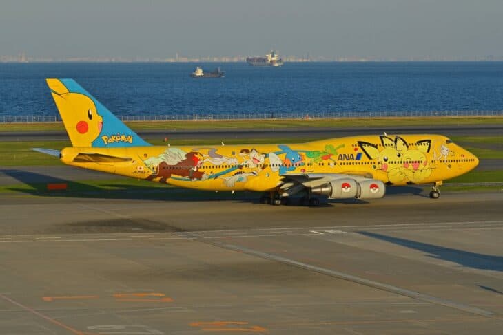 All-yellow ANA Boeing 747-400D JA8957 in the Pikachu Jumbo livery taxiing at Tokyo Haneda, with Tokyo Bay and shipping behind it.