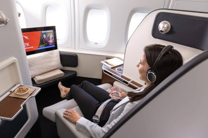 Passenger with headphones reclining in a Qantas A380 first class suite, with a grey leather seat and a large screen facing her