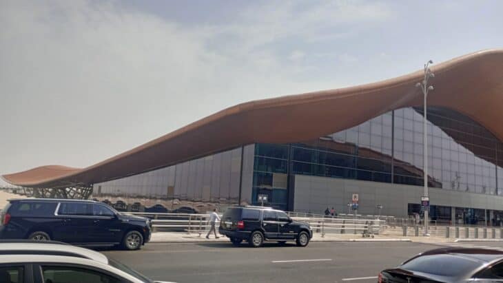 The curved, wave-shaped roof and glass facade of a passenger terminal at King Abdulaziz International Airport in Jeddah, with SUVs on the forecourt