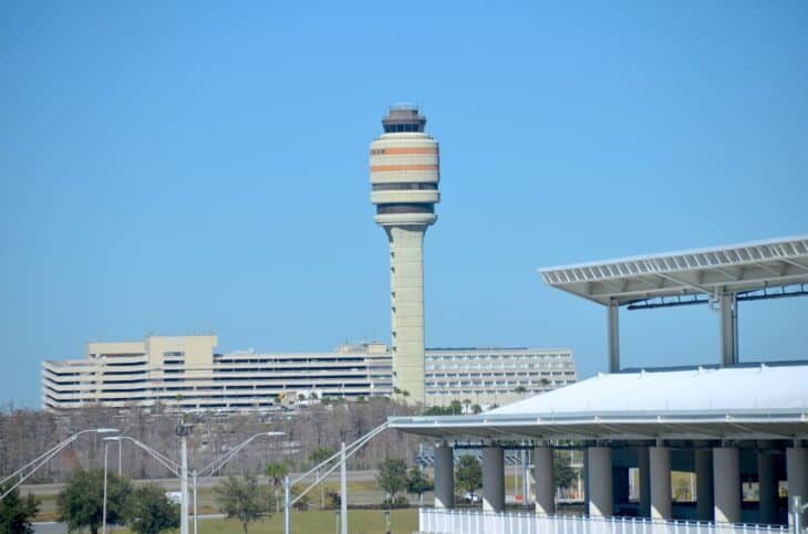 The tall striped air traffic control tower at Orlando International Airport against a blue sky, with terminal buildings and a white canopy structure below