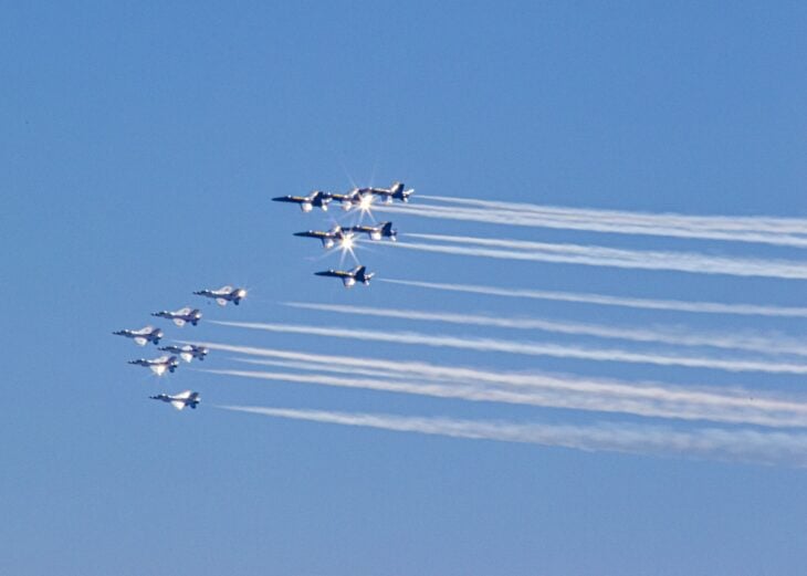 Six Blue Angels F/A-18 Hornets flying in formation above six Air Force Thunderbirds F-16s, both trailing smoke across a blue sky