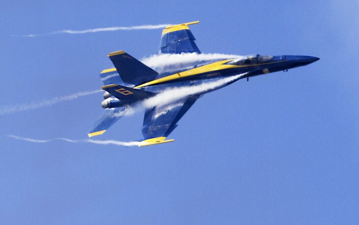 A blue and gold Blue Angels F/A-18 Super Hornet banking hard with condensation streaming off its wings
