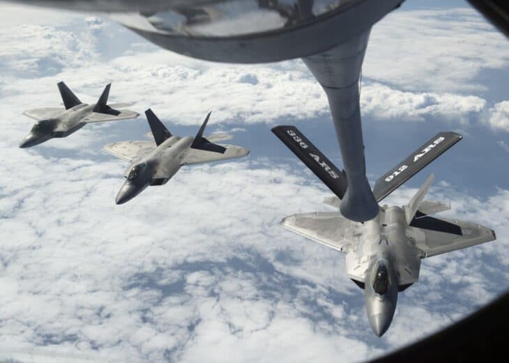 Three F-22 Raptors in echelon behind the extended KC-135R refueling boom above white clouds, seen from the boom pod window