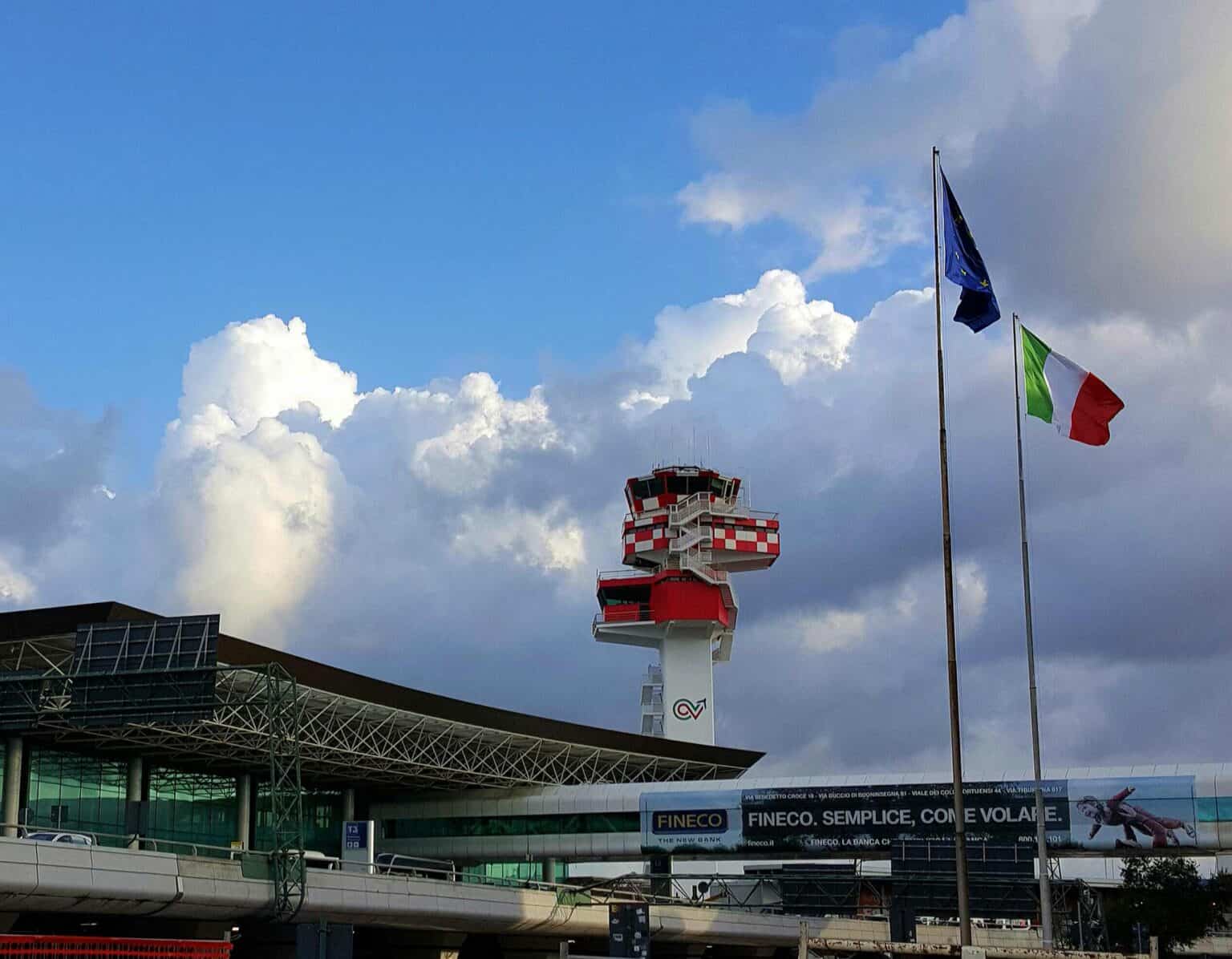 Red-and-white ENAV air traffic control tower at Rome Fiumicino Airport behind the terminal, with the Italian and EU flags flying