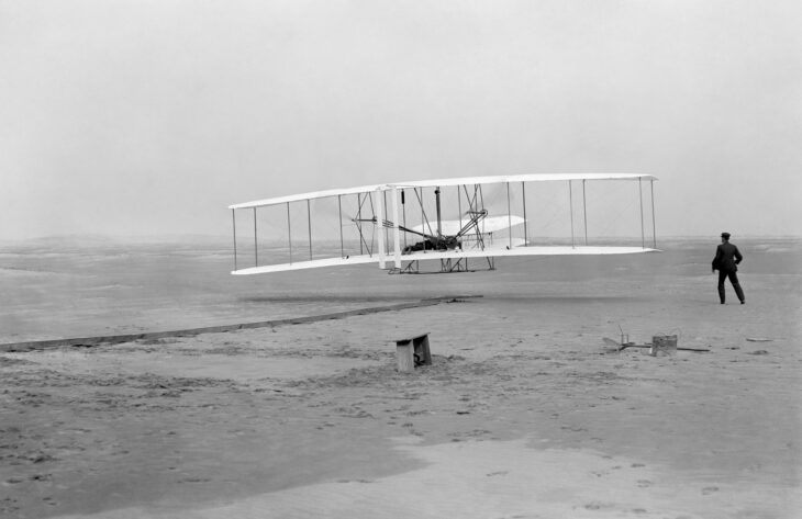 The Wright Flyer lifting off the sand at Kill Devil Hills on December 17, 1903, with Wilbur Wright standing at right