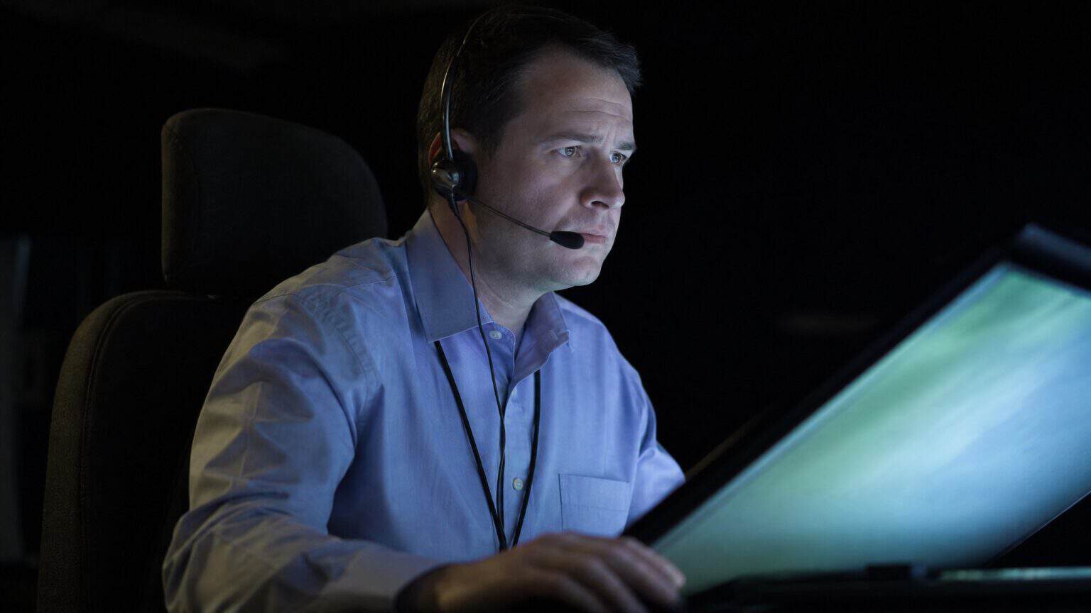 An air traffic controller wearing a headset works at a radar console in a darkened control room, face lit by the glow of the display